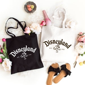 May include: Two canvas tote bags, one black with white text and one white with black text. Both bags feature the word Disneyland in a stylized font, the text EST. 1955, and a graphic of Mickey Mouse. The bags have two shoulder straps each.