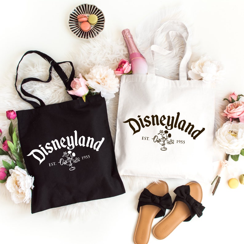 May include: Two canvas tote bags, one black with white text and one white with black text. Both bags feature the word Disneyland in a stylized font, the text EST. 1955, and a graphic of Mickey Mouse. The bags have two shoulder straps each.