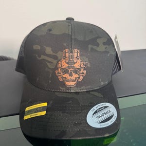 Peut inclure: Une casquette de camionneur camouflage noir et vert avec un crâne portant un casque et des lunettes de vision nocturne. La casquette a un dos en maille noire et un autocollant jaune qui dit "MULTICAM BLACK". La casquette a également un autocollant blanc qui dit "THE AUTHENTIC SNAPBACK YUPOONG SINCE 1974".