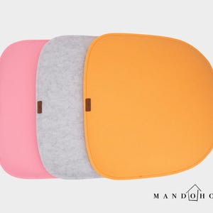 May include: Three felt placemats in pink, gray, and orange. Each placemat has a rounded square shape with a small brown accent. The brand name "MANDO HOME" is printed in black at the bottom.