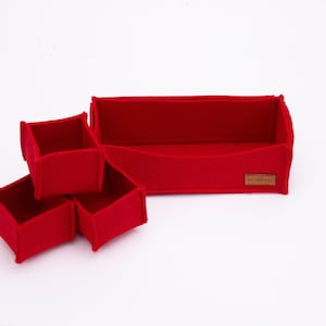 May include: A set of bright red felt storage organizers consisting of one rectangular tray and three smaller square containers. The rectangular tray features a small brown leather-like brand tag on the front right side with visible text.