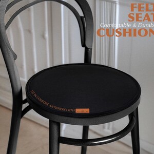 Peut inclure: Un coussin d'assise rond en feutre noir posé sur une chaise en bois noir. Le coussin porte le texte FELT SEAT Comfortable & Durable CUSHION et SIT IN COMFORT, ANYWHERE WITH, ainsi qu'une petite étiquette de marque rectangulaire marron.