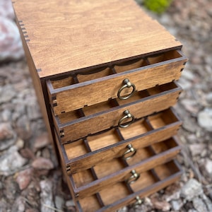 Può includere: Un portagioie in legno con cinque cassetti, ciascuno con una piccola maniglia rotonda in metallo. La scatola è tinta di un caldo colore marrone e i cassetti sono aperti, rivelando piccoli scomparti. La scatola è appoggiata su pacciame marrone.