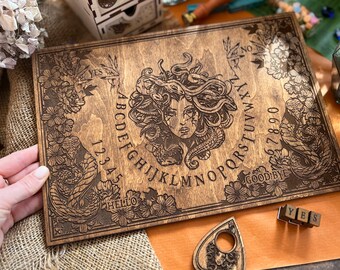 May include: A wooden Ouija board with a dark brown stain. The board features a Medusa illustration with snakes and floral accents. The letters, numbers, and words "YES", "NO", "GOODBYE", and "HELLO" are printed on the board.