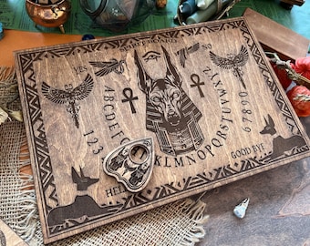 May include: A wooden spirit board with an Anubis design, featuring an alphabet, numbers, yes, no, and goodbye. A planchette is included.