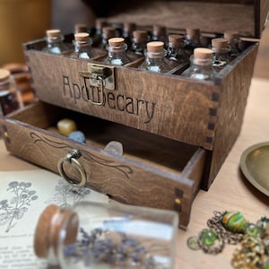 May include: Apothecary wooden box with a drawer, filled with small glass bottles with cork stoppers. The word "Apothecary" is engraved on the front. The drawer contains small crystals. A vintage aesthetic.