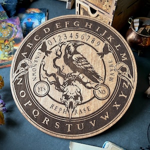 Puede incluir: Tablero de Ouija de madera con un diseño de cuervo y calavera. El tablero presenta el alfabeto, los números del 0 al 9, sí, no, tal vez, desconocido y reformular.