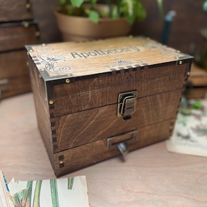Puede incluir: Una caja de boticario de madera marrón oscuro con esquinas metálicas y un cierre. La palabra "Apothecary" está grabada en la tapa. La caja tiene un cajón y probablemente se utiliza para guardar hierbas u otros artículos. Mide unos 20 cm de alto.