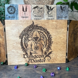 Puede incluir: Una caja de madera con tapa con bisagras, que presenta un dragón y el texto "Dungeon Master". La caja tiene seis compartimentos, cada uno etiquetado con un tipo de madera diferente: Negro, Cerezo, Nogal Medio, Nogal Oscuro y Chocolate.