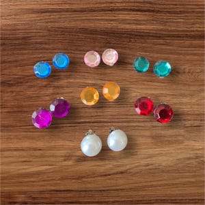 May include: A collection of colorful clip-on earrings in various jewel tones, including blue, pink, green, orange, red, purple, and white. The earrings are arranged on a wooden surface, showcasing their faceted designs and pearl-like finishes.
