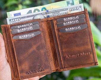 May include: A brown leather wallet with multiple card slots and a personalized inscription that reads "Ahmet Mert". The wallet is open and has several cards inside, including a $100 bill.