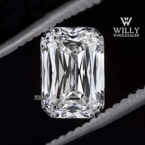 May include: A clear, rectangular, emerald cut diamond gemstone held by a pair of tweezers. The diamond is reflecting light and has a brilliant shine. Willy Wholesaler is printed in white text on a black background.