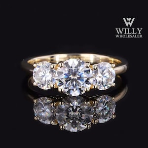 May include: A gold band ring with three large, round, clear gemstones. The ring is set against a black background, with a reflection of the ring visible below. The text "Willy Wholesaler" is in the upper right corner.