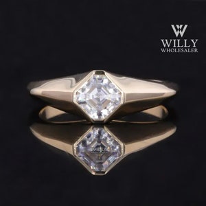 May include: A gold ring with a square-shaped, clear gemstone set in a bezel setting. The ring has a thick band with a slightly rounded top.