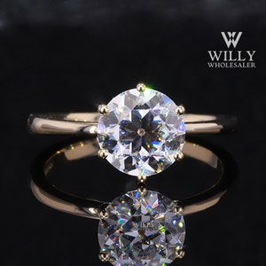 Puede incluir: Un anillo de compromiso dorado con una gran piedra preciosa redonda y transparente. El anillo está engastado con seis garras y refleja la luz, creando un efecto brillante. El anillo se muestra sobre una superficie negra reflectante.