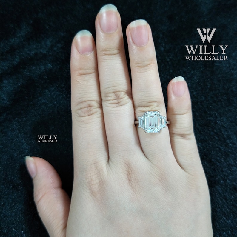 May include: A silver ring with a large emerald-cut center stone and two smaller emerald-cut stones on either side. The ring is on a person's finger.