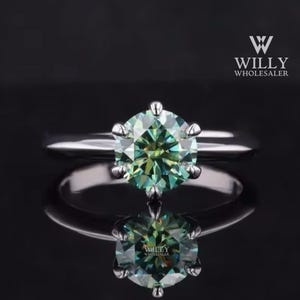 Peut inclure: Une bague en argent avec une grosse pierre de moissanite ronde, verte et bleue. La bague est posée sur un fond noir.