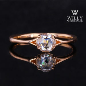 Puede incluir: Anillo de oro rosa con una gema grande, clara y facetada. El anillo tiene una banda delgada y un engaste distintivo con detalles en naranja. El anillo se muestra sobre una superficie negra reflectante, con el logotipo "WILLY WHOLESALER".
