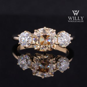 Op de afbeelding: Een gouden ring met drie stenen. De middelste steen is een grote, vierkant geslepen, champagnekleurige diamant. De twee zijstenen zijn kleinere, ronde, witte diamanten.
