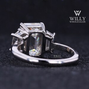 May include: A silver ring with a large emerald-cut center stone and two smaller stones on either side. The ring is set in a prong setting and has a polished finish.