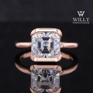 Può includere: Anello in oro rosa con una grande gemma trasparente a taglio quadrato. La gemma è incastonata in una lunetta. L'anello è esposto su una superficie nera riflettente, con il testo "WILLY WHOLESALER".