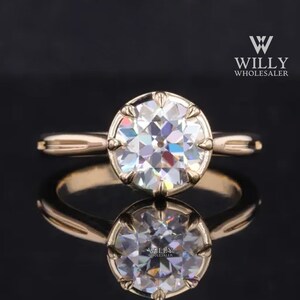 Puede incluir: Un anillo de compromiso de oro con un gran diamante redondo y transparente. El diamante tiene un corte facetado y está engastado en un engaste de garras. La banda del anillo es fina y tiene un diseño simple.