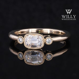 May include: A rose gold ring with a large, rectangular, clear gemstone in the center, flanked by two smaller, round, clear gemstones. The ring has a delicate, beaded bezel setting and is displayed on a reflective surface.