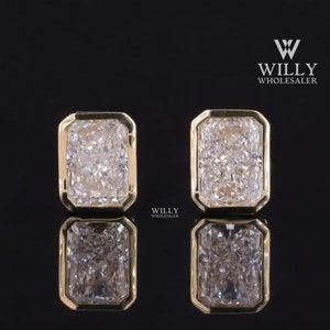 Op de afbeelding: Een paar rechthoekige diamanten oorstekers met een gouden bezel-zetting. De diamanten zijn helder en sprankelend en reflecteren licht. De oorbellen worden gepresenteerd op een reflecterend zwart oppervlak.