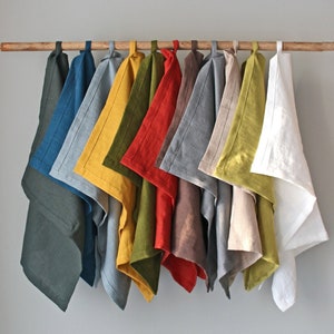 May include: A set of 12 linen tea towels in various colors, including teal, gray, yellow, green, red, gray, beige, and white. The towels are hanging on a wooden rod.
