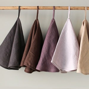 May include: Five linen tea towels hanging on a wooden rod. The towels are in shades of gray, brown, purple, pink, and beige.