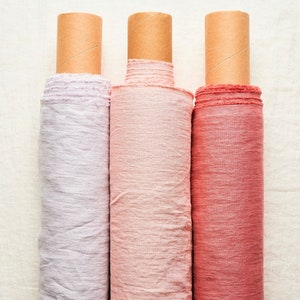 May include: Three rolls of fabric in different shades of pink, from light to dark, are rolled up on cardboard tubes. The fabric is a linen blend and has a slightly textured surface.