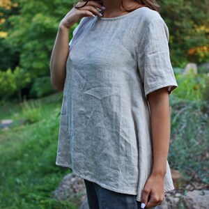 May include: A light beige linen top with a round neckline and short sleeves. The top has a relaxed fit and a slightly A-line shape.