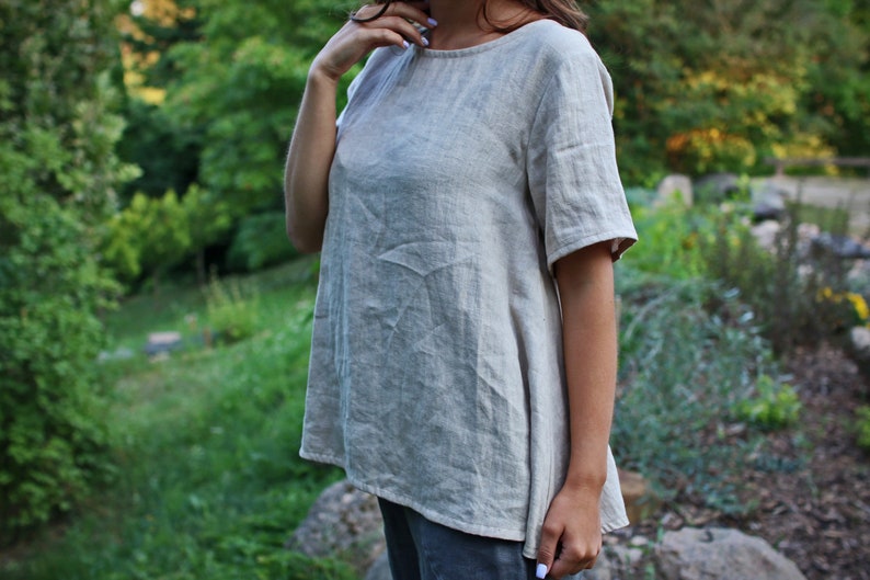 May include: A light beige linen top with a round neckline and short sleeves. The top has a relaxed fit and a slightly A-line shape.