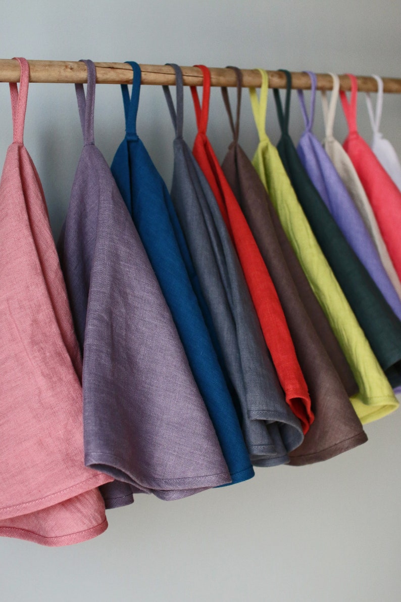 May include: A set of 12 linen tea towels hanging on a wooden rod. The towels are in various colors, including pink, purple, blue, gray, red, brown, yellow, green, and lavender.