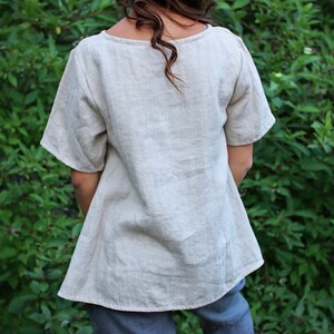 May include: A light beige linen top with a scoop neck and short sleeves. The top is loose fitting and has a slightly A-line shape.