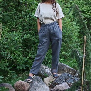 May include: A person wearing a light beige linen top and dark gray linen pants with a drawstring waist. The person is standing on rocks in a garden setting.