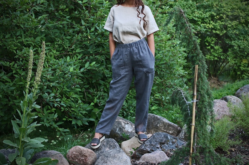 May include: A person wearing a light beige linen top and dark gray linen pants with a drawstring waist. The person is standing on rocks in a garden setting.