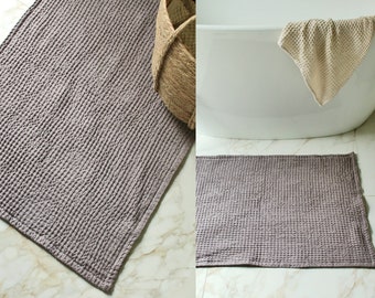 May include: Two gray waffle-weave bath mats, one on a white tiled floor and the other in front of a white bathtub. The bath mat is made of a soft, absorbent material.