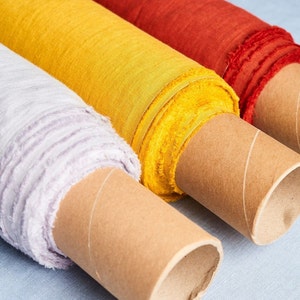 Pode incluir: Três rolos de tecido, um amarelo, um vermelho e um roxo claro, estão enrolados em tubos de cartão. O tecido é texturizado e tem uma borda ligeiramente desfiada.