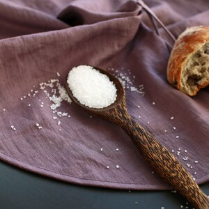 May include: A wooden spoon filled with white salt crystals sits on a purple linen cloth. The spoon has a dark brown wood grain pattern.
