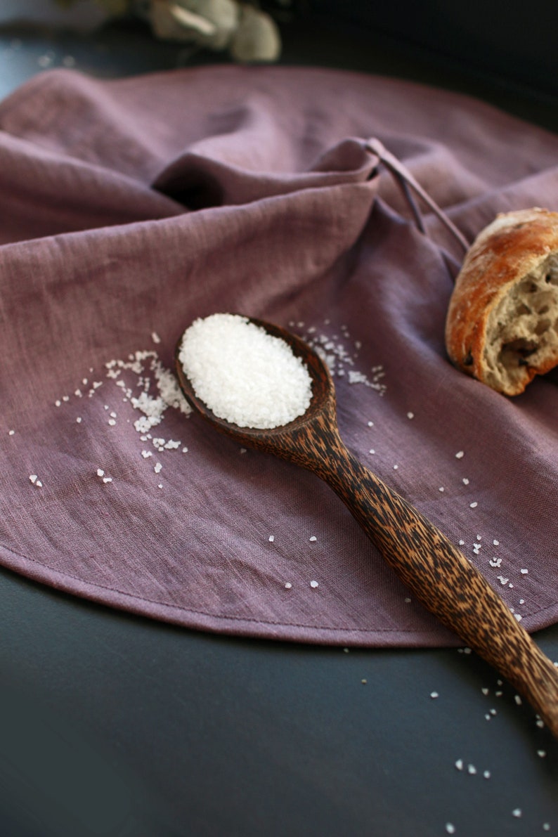 May include: A wooden spoon filled with white salt crystals sits on a purple linen cloth. The spoon has a dark brown wood grain pattern.