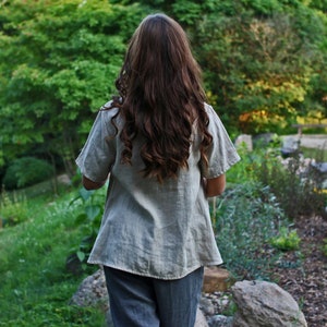 May include: A woman wearing a light gray linen shirt with short sleeves and gray pants. She is standing in a garden with green trees and plants in the background.