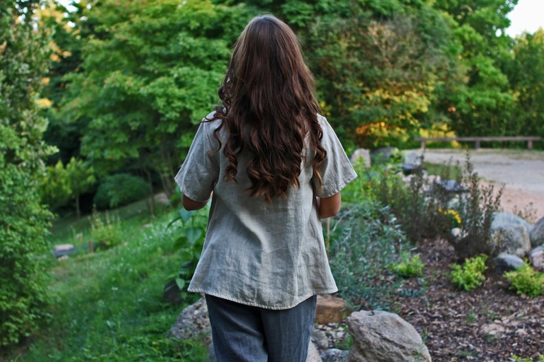 May include: A woman wearing a light gray linen shirt with short sleeves and gray pants. She is standing in a garden with green trees and plants in the background.