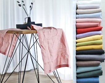 May include: A stack of 14 linen tablecloths in various colors, including white, beige, light blue, pink, red, lavender, yellow, green, dark gray, light gray, brown, teal, and blue. The tablecloths are folded and stacked on a white shelf. A pink linen tablecloth is draped over a black metal table with a wooden top.
