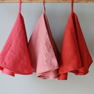 May include: Three linen tea towels hanging on a wooden rod. The towels are in shades of red, pink, and coral.