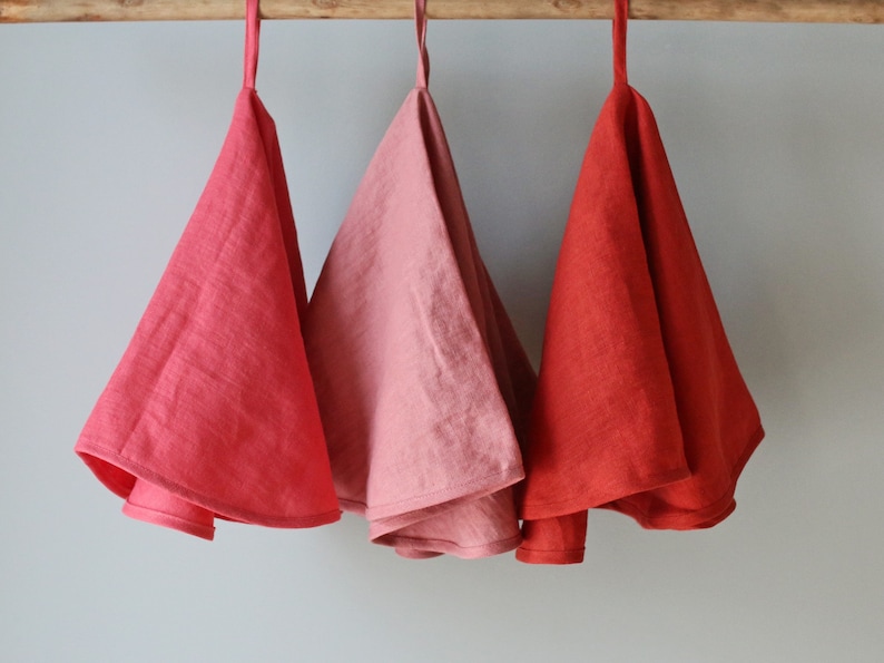 May include: Three linen tea towels hanging on a wooden rod. The towels are in shades of red, pink, and coral.