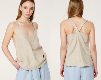 May include: A beige linen tank top with a V-neck and criss-cross straps in the back. The model is wearing the tank top with light blue shorts.