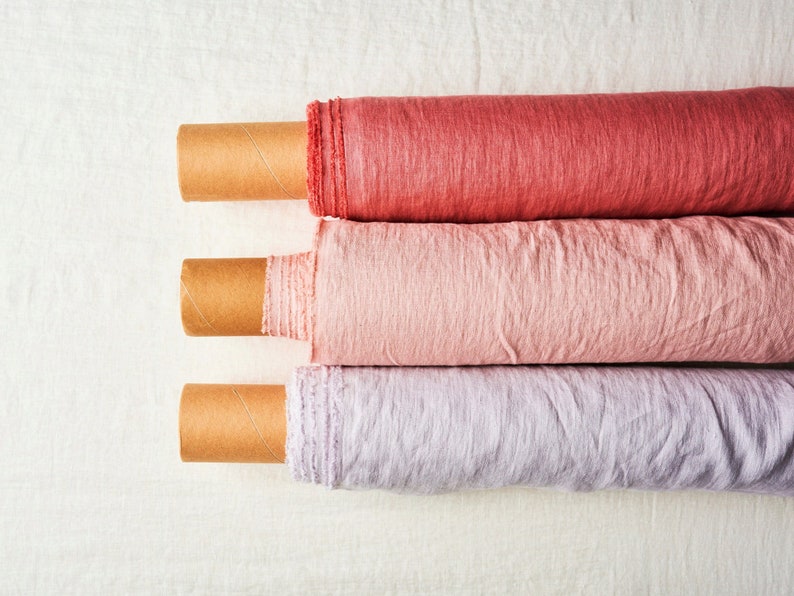 May include: Three rolls of fabric in different shades of pink and purple. The fabric is rolled up and secured with cardboard tubes. The fabric is a linen blend.