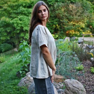 May include: A woman wearing a light beige linen top with short sleeves and a scoop neck. She is standing in a garden setting with green foliage and rocks.