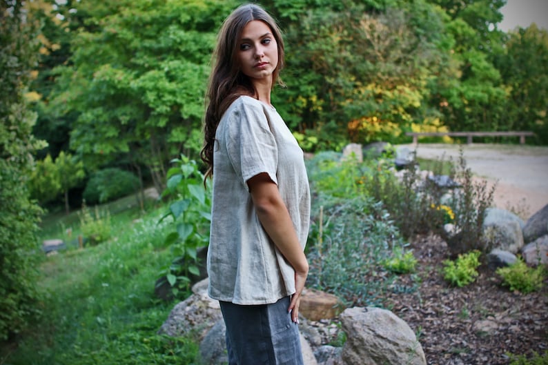 May include: A woman wearing a light beige linen top with short sleeves and a scoop neck. She is standing in a garden setting with green foliage and rocks.